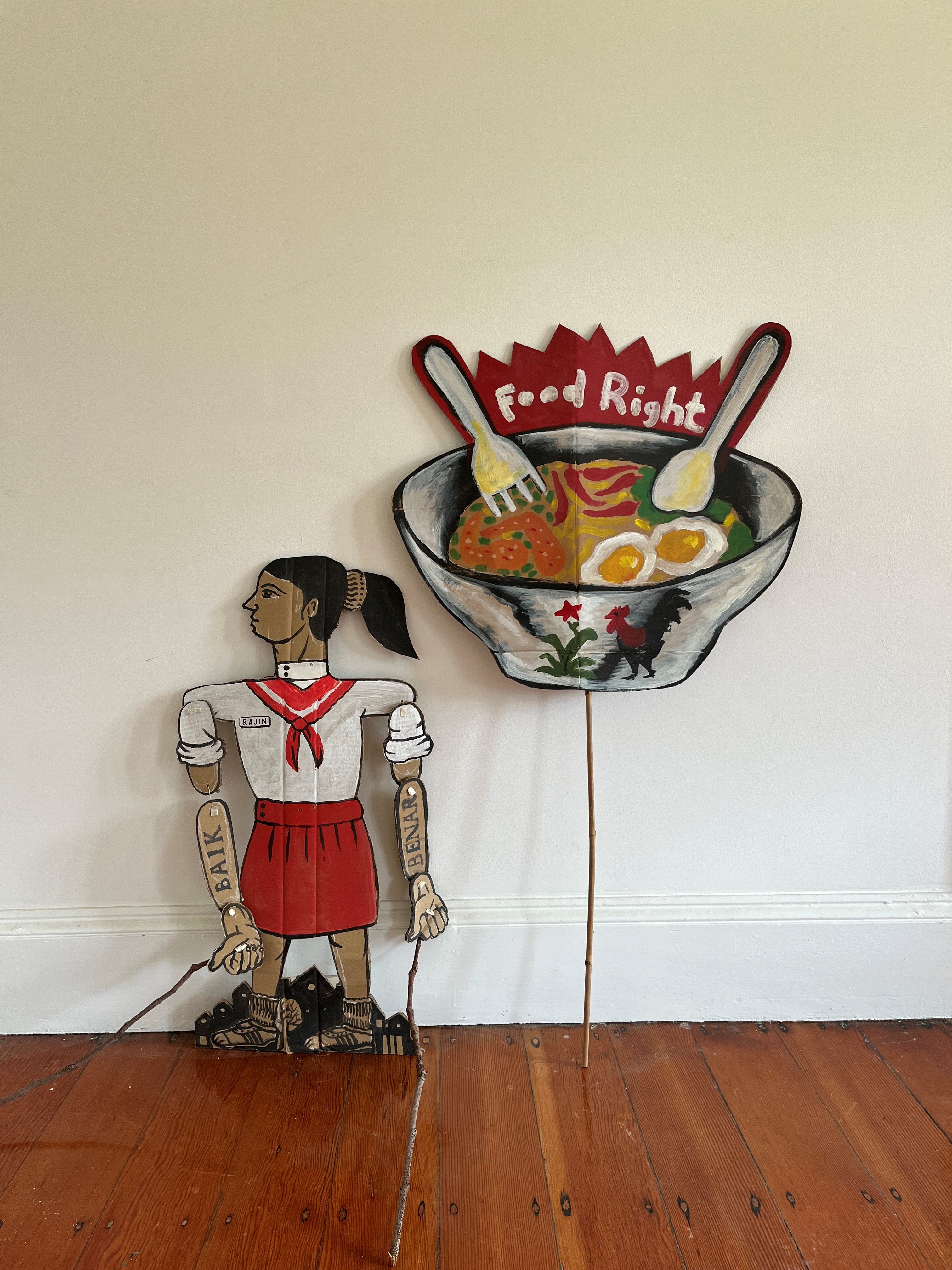 Artist Deni stands next to artwork Food right, a puppet of a girl in uniform standing above a bowl of food with the words food right written on the bowl, artist Safdar Ahmed stands next to him